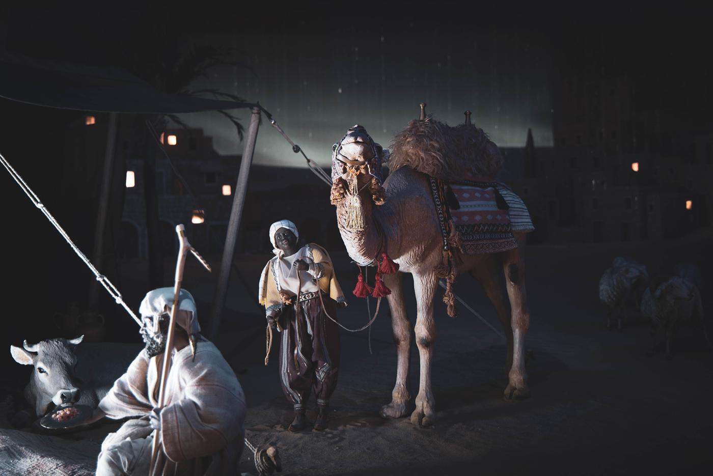 Il presepio del Natale 2022, di notte: sotto una tenda, un pastore seduto con il bastone e un bue; al centro un ragazzo con il turbante tiene un cammello bardato; dietro, le finestre accese del villaggio.